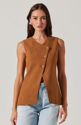 A woman is wearing a sleeveless, camel-colored vest with a V-neckline. The vest has three large, round buttons running diagonally across the front. It has a tailored fit and extends slightly below the waist. She pairs it with light blue, high-waisted jeans.